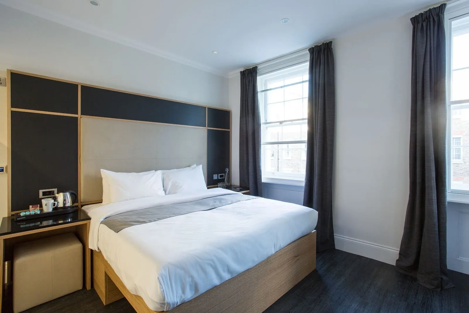 A room at the Z Hotel Gloucester Place with a double bed on a pale wooden base, a black and oak panelled headboard, a kettle on the built-in side shelf and two tall sash windows with charcoal curtains