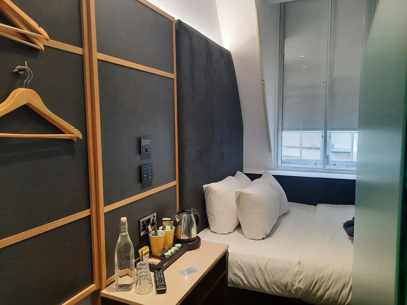 A room at the Z Hotel Holborn with a dark upholstered headboard, a kettle and glasses on the desk, and a window with the blind partly raised