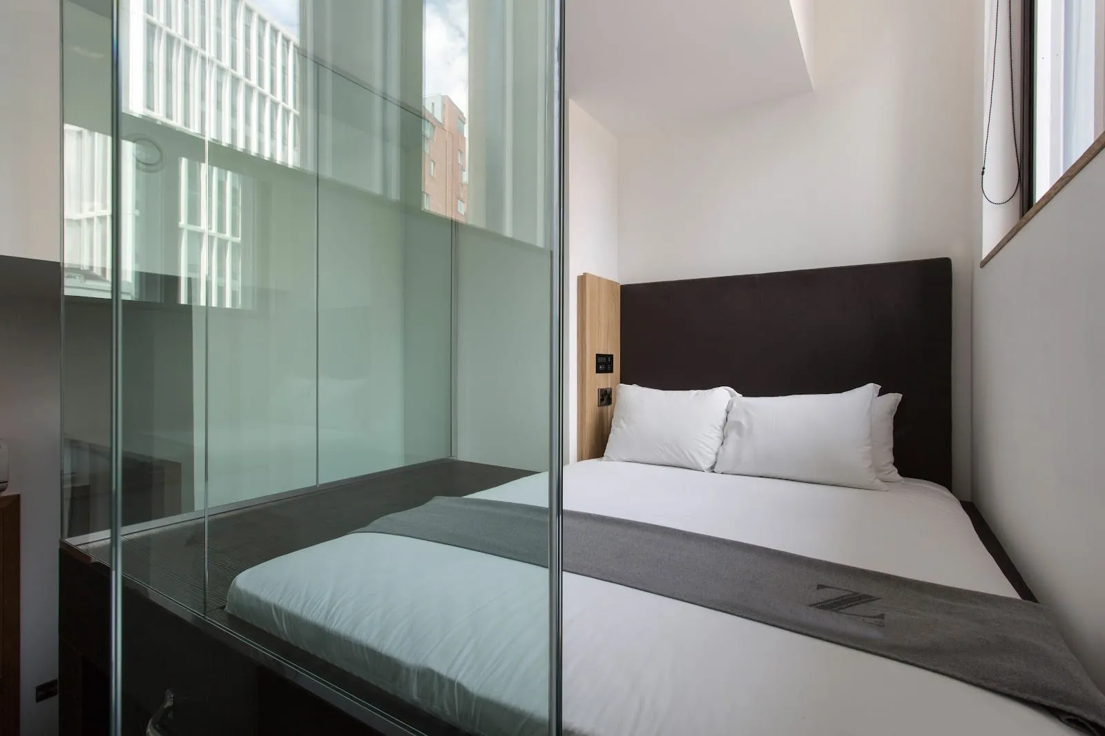 A room at the Z Hotel Shoreditch with a dark upholstered headboard, a glass-walled shower cubicle and a window looking onto neighbouring buildings