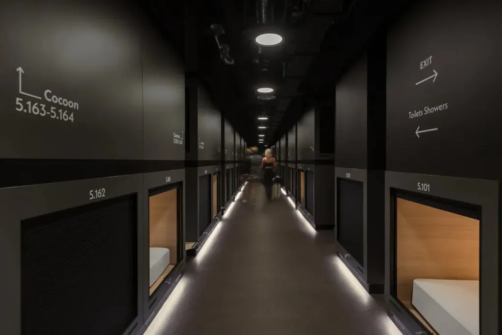 A corridor of sleeping capsules at a Zedwell capsule hotel, lit strips along the floor and signage reading Cocoon 5.163-5.164 and Toilets Showers