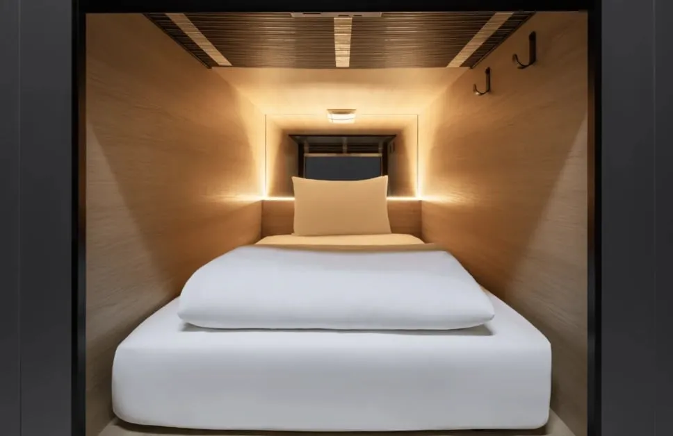 Inside a sleeping capsule, a mattress and pillow in a wood-lined berth with hooks and a shelf, too low to stand in