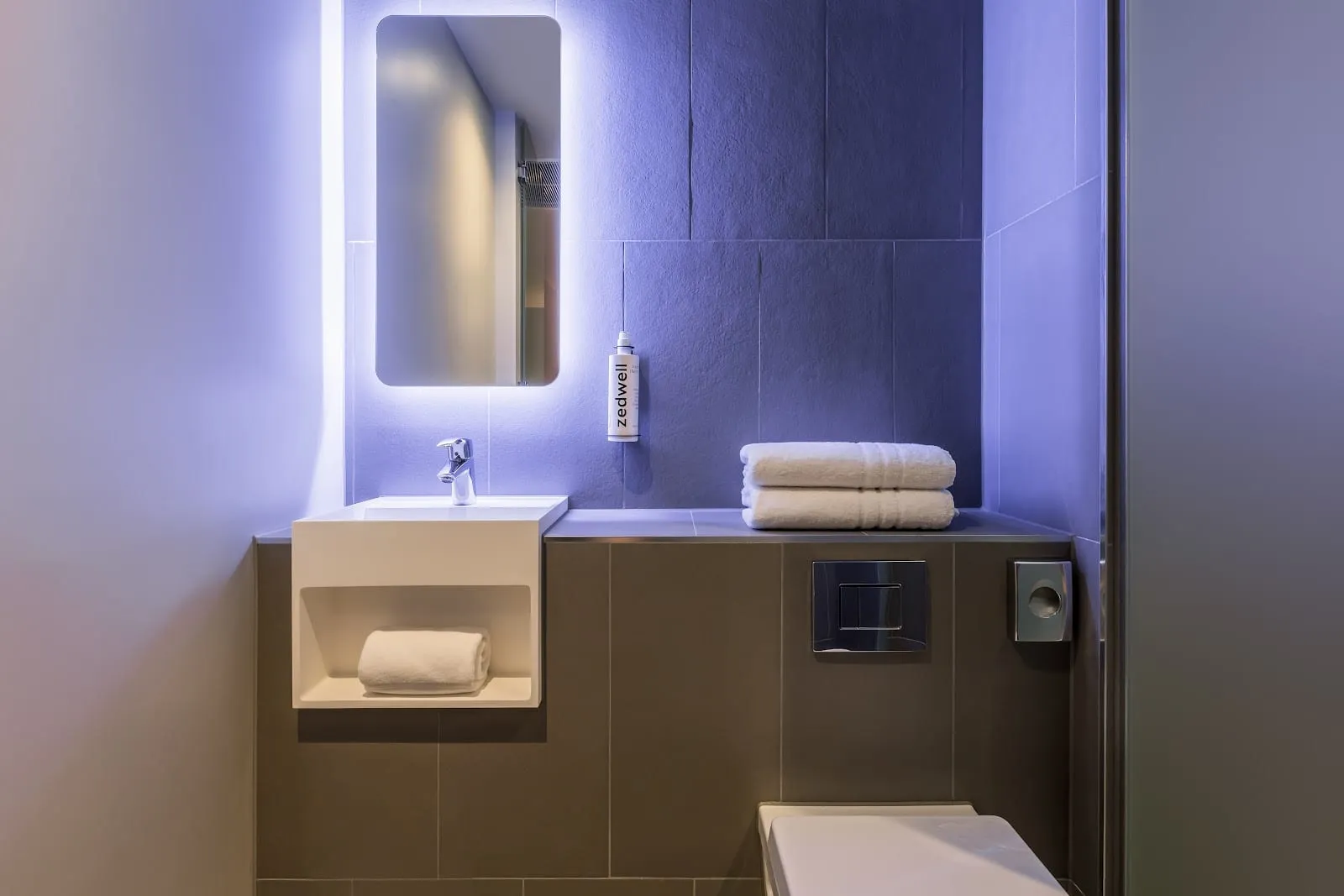 The ensuite bathroom of a Zedwell Greenwich room, with a backlit mirror, folded towels and Zedwell-branded amenities
