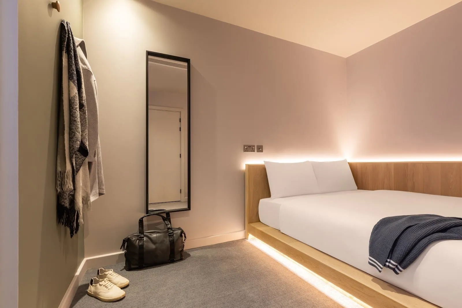 A windowless Cocoon room at Zedwell Greenwich: a low oak platform bed with warm LED strips under the base and along the headboard, pale pink walls, a tall black-framed mirror and coat hooks