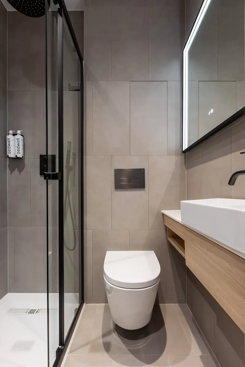 The ensuite bathroom of a Zedwell Knightsbridge room, with a walk-in glass shower, a floating wood vanity and Zedwell-branded amenities