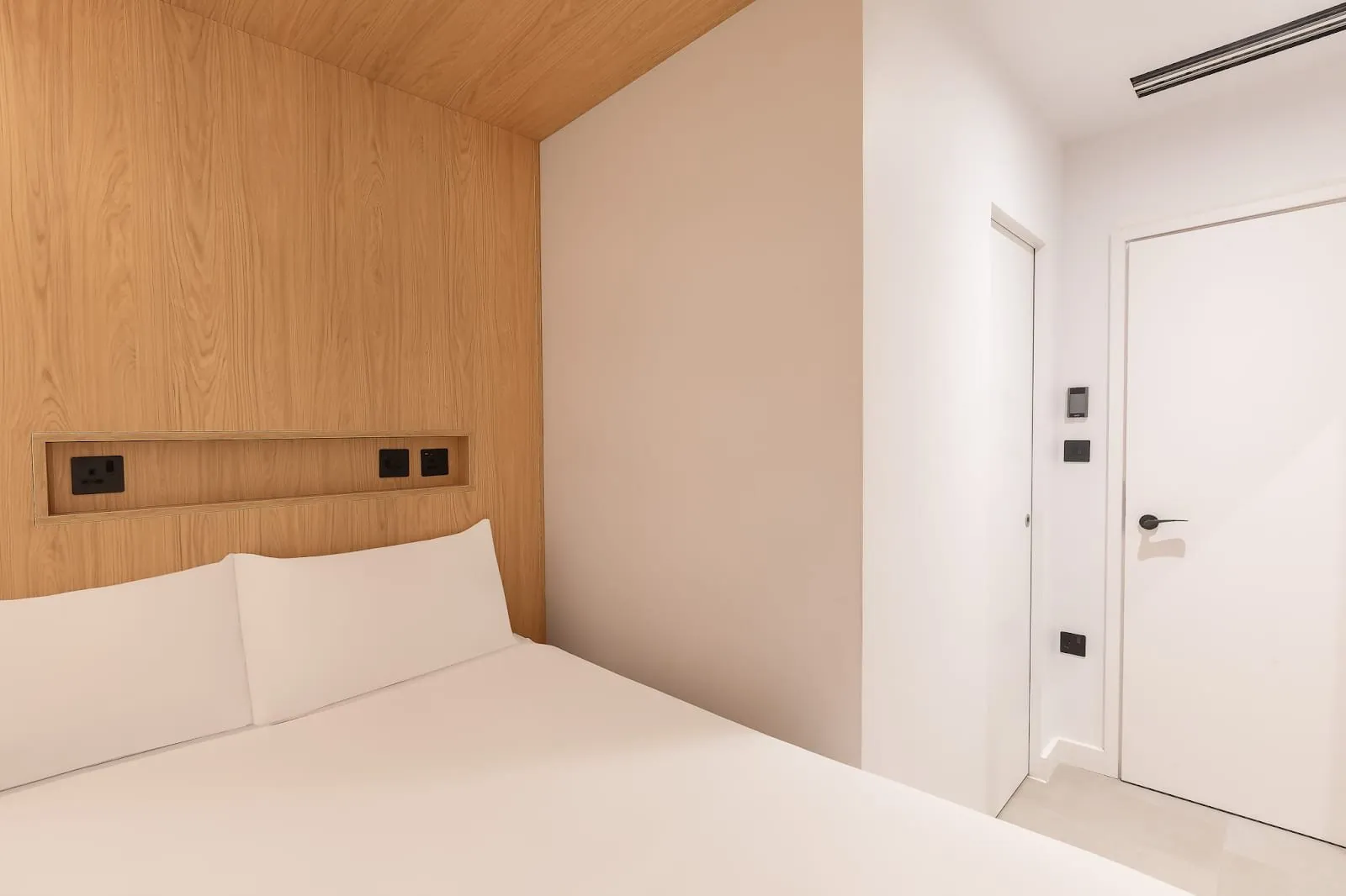 A Cocoon 2 room at Zedwell Knightsbridge: a double bed against an oak-panelled wall with built-in sockets, white walls with no window, a sliding door to the shower room and the door to the corridor