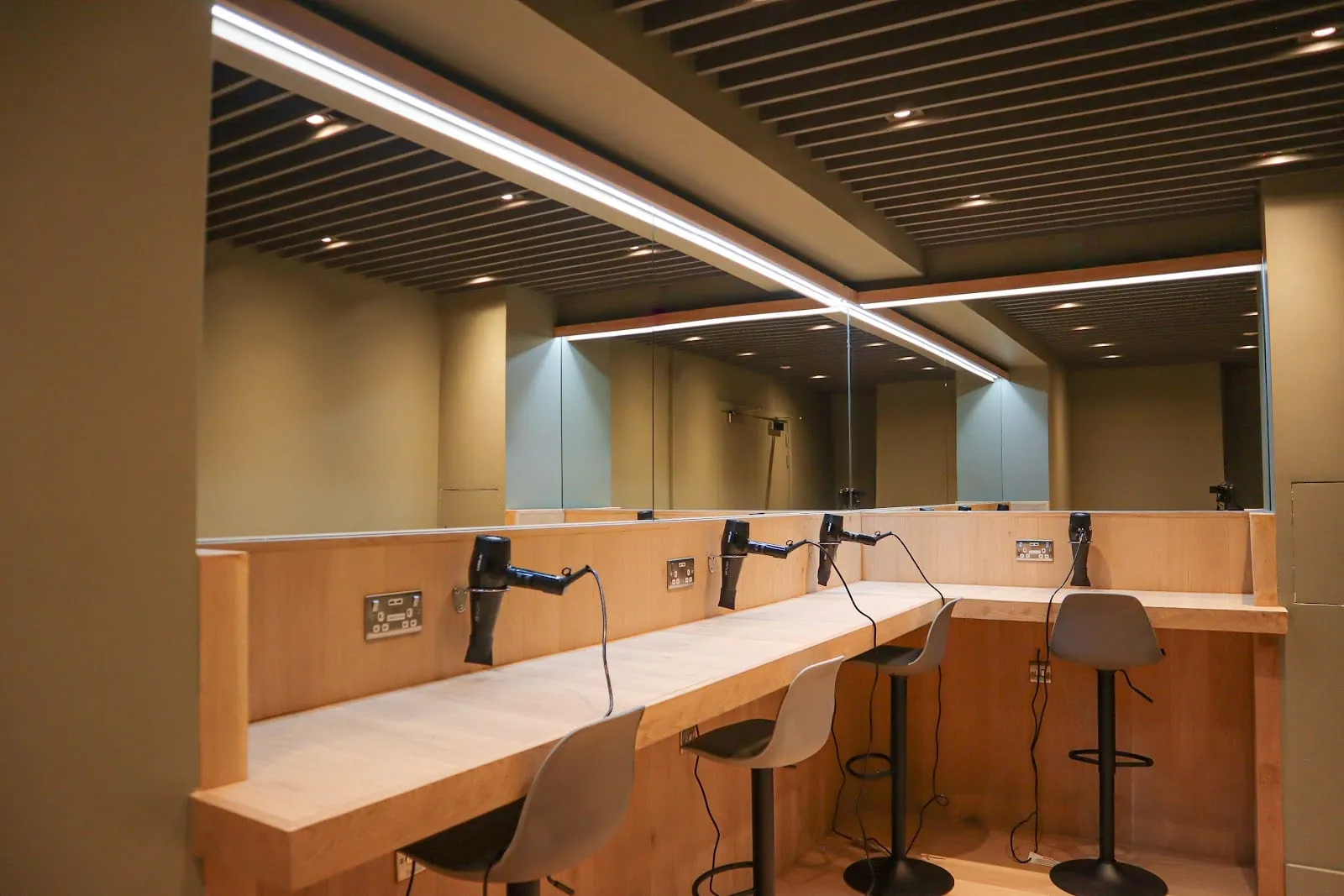A row of vanity stations with wall-mounted hairdryers and stools facing a long mirror, with a shower stall visible behind the glass partition