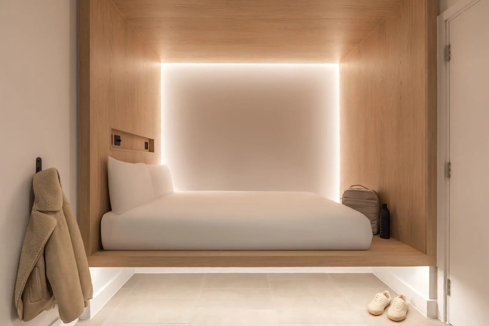 A Zedwell Park Lane Cocoon: a double mattress on a raised oak platform set into a backlit oak alcove, a coat on a wall hook and a pale tiled floor, with no window