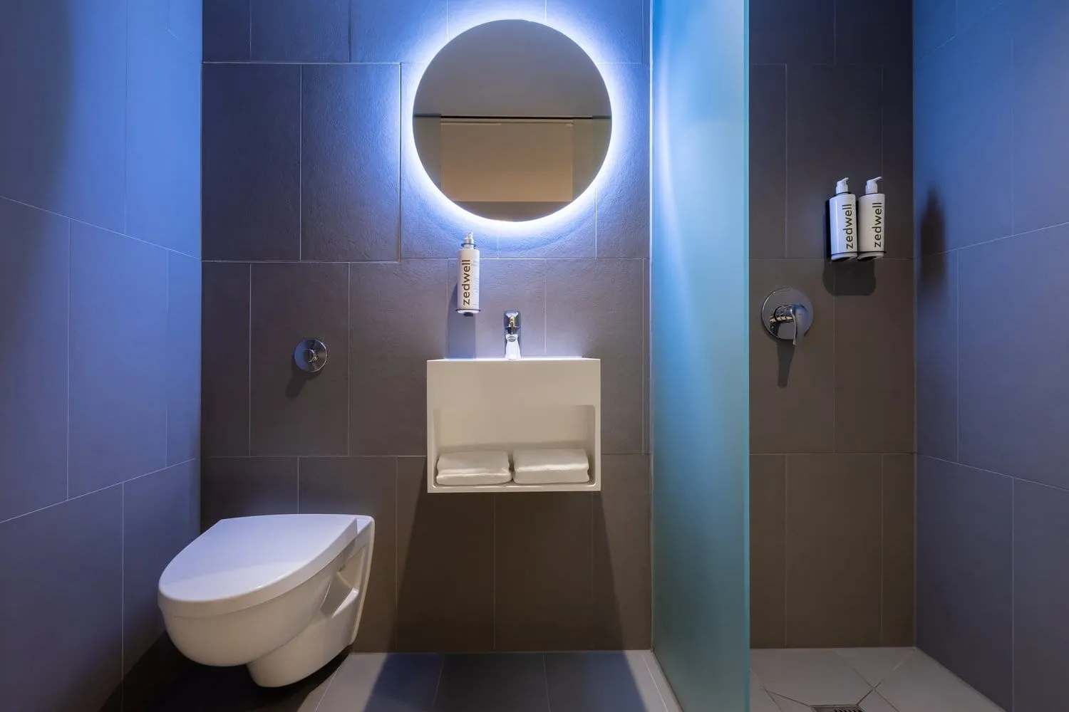 The ensuite bathroom of a Zedwell Tottenham Court Road room, with a round backlit mirror, Zedwell-branded amenities and a walk-in shower