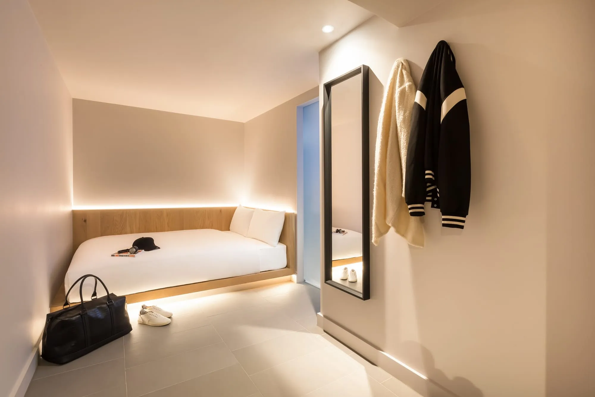 A Cocoon room at Zedwell Tottenham Court Road, with the bed on a wood platform and a jacket hanging beside a full-length mirror